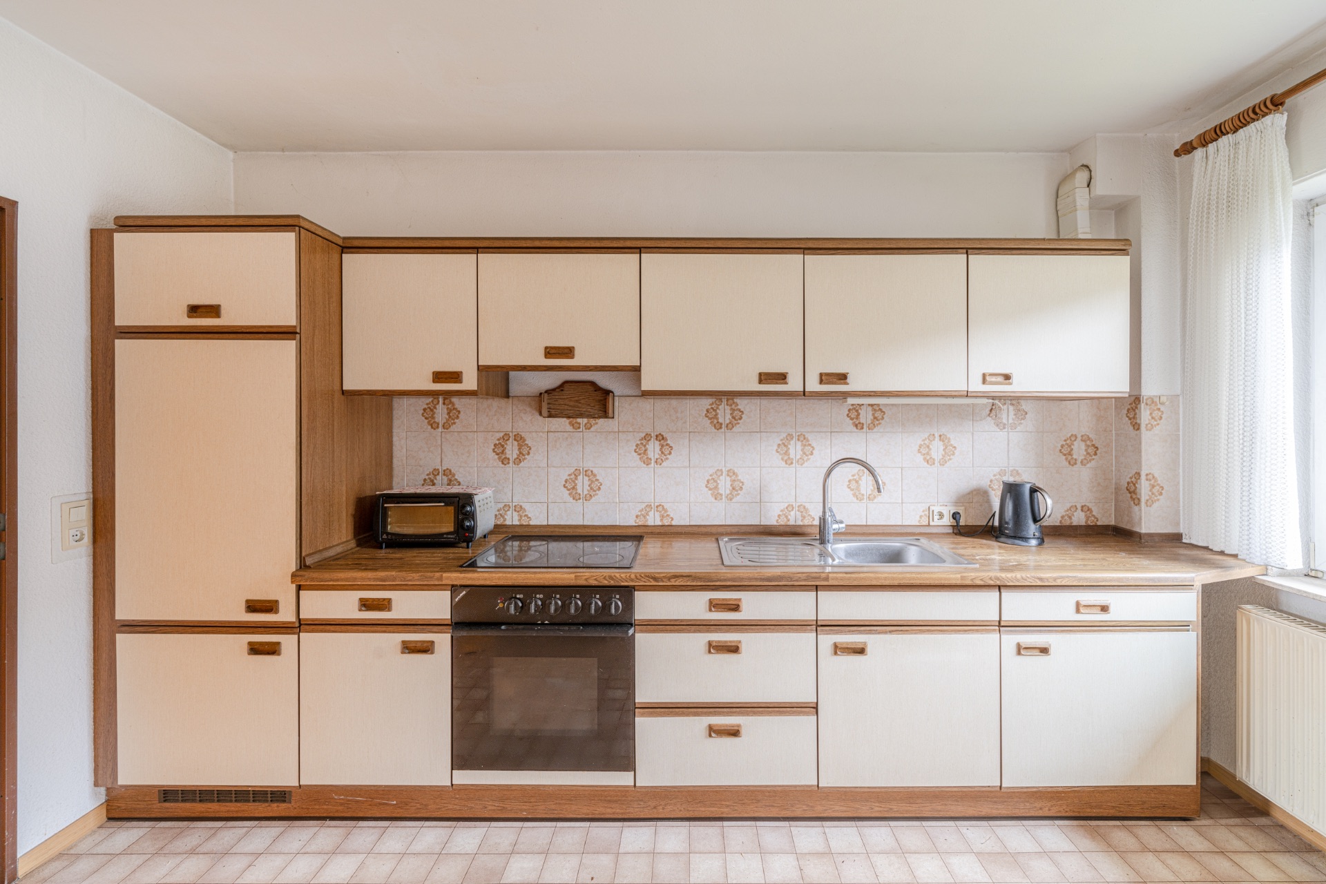 Küchenzeile mit hellen Schränken, Backofen, Spüle, Wasserkocher und Toaster vor gemusterter Fliesenwand.