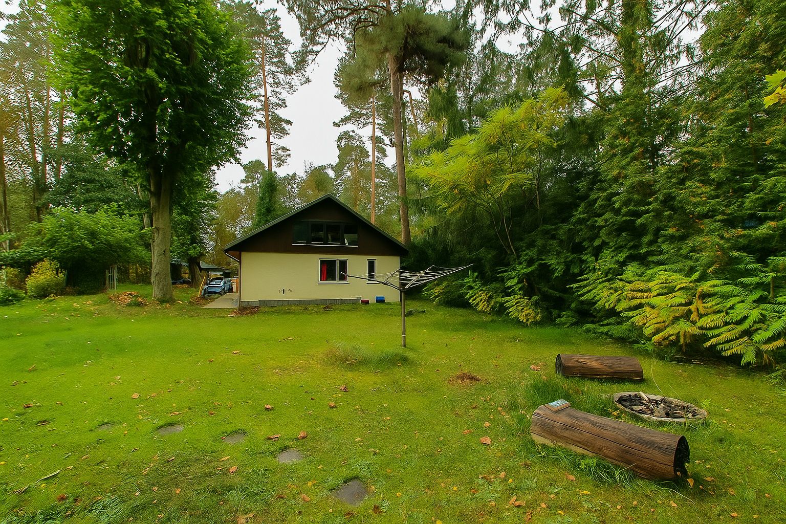 Haus mit Satteldach in grünem Garten, Feuerstelle und Baumstämmen im Vordergrund.