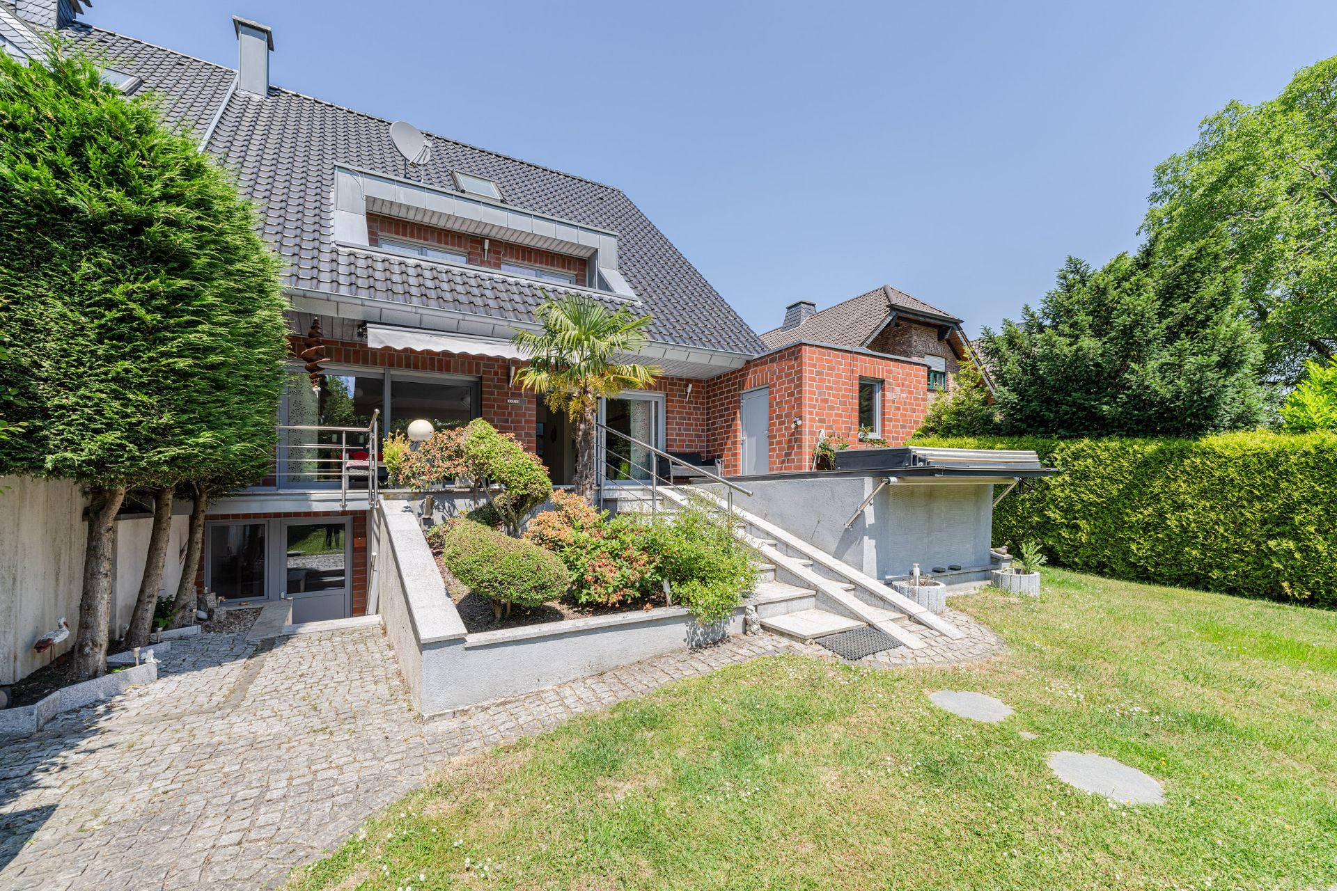 Modernes Backsteinhaus mit Garten, Terrasse, Rampe und gepflegten Büschen im Vordergrund.
