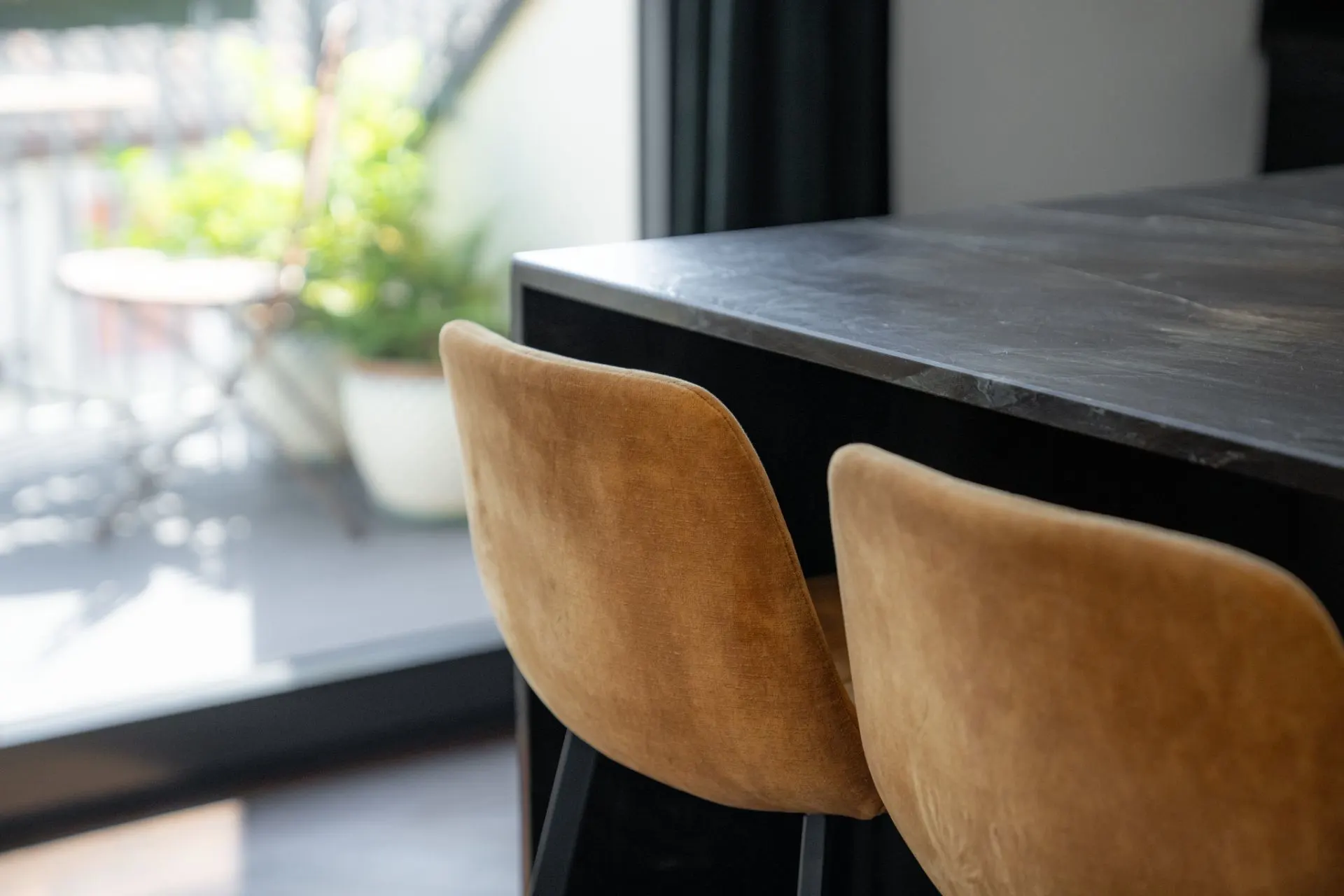 Zwei braune Stühle stehen an einem dunklen Tisch, im Hintergrund ein Balkon mit Pflanzen.