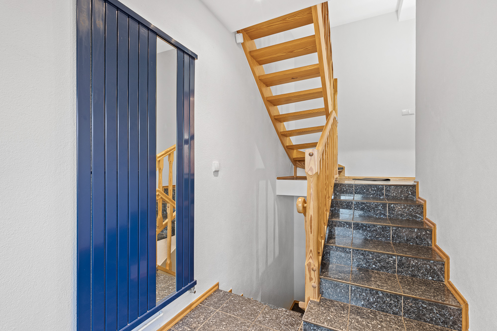 Treppenhaus mit Holzgeländer, dunklen Fliesen und großem blauen Spiegel an der Wand.