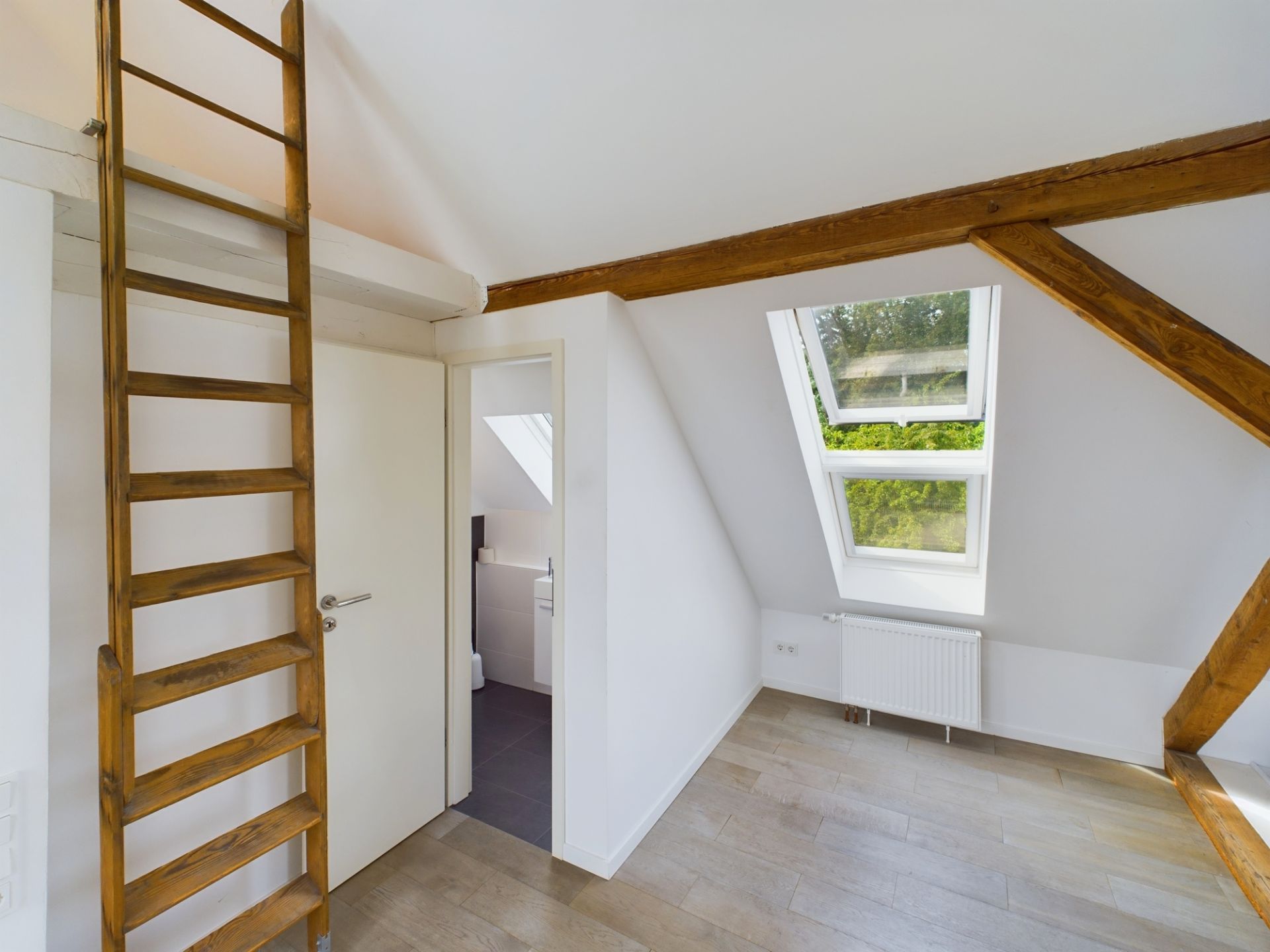 Helles Zimmer mit Holzleiter, Dachfenstern, Holzbalken und Blick ins kleine Bad.