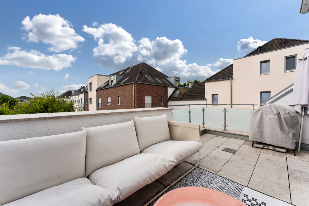 Moderne Terrasse mit weißem Sofa, Grill und Blick auf Wohngebäude unter blauem Himmel.