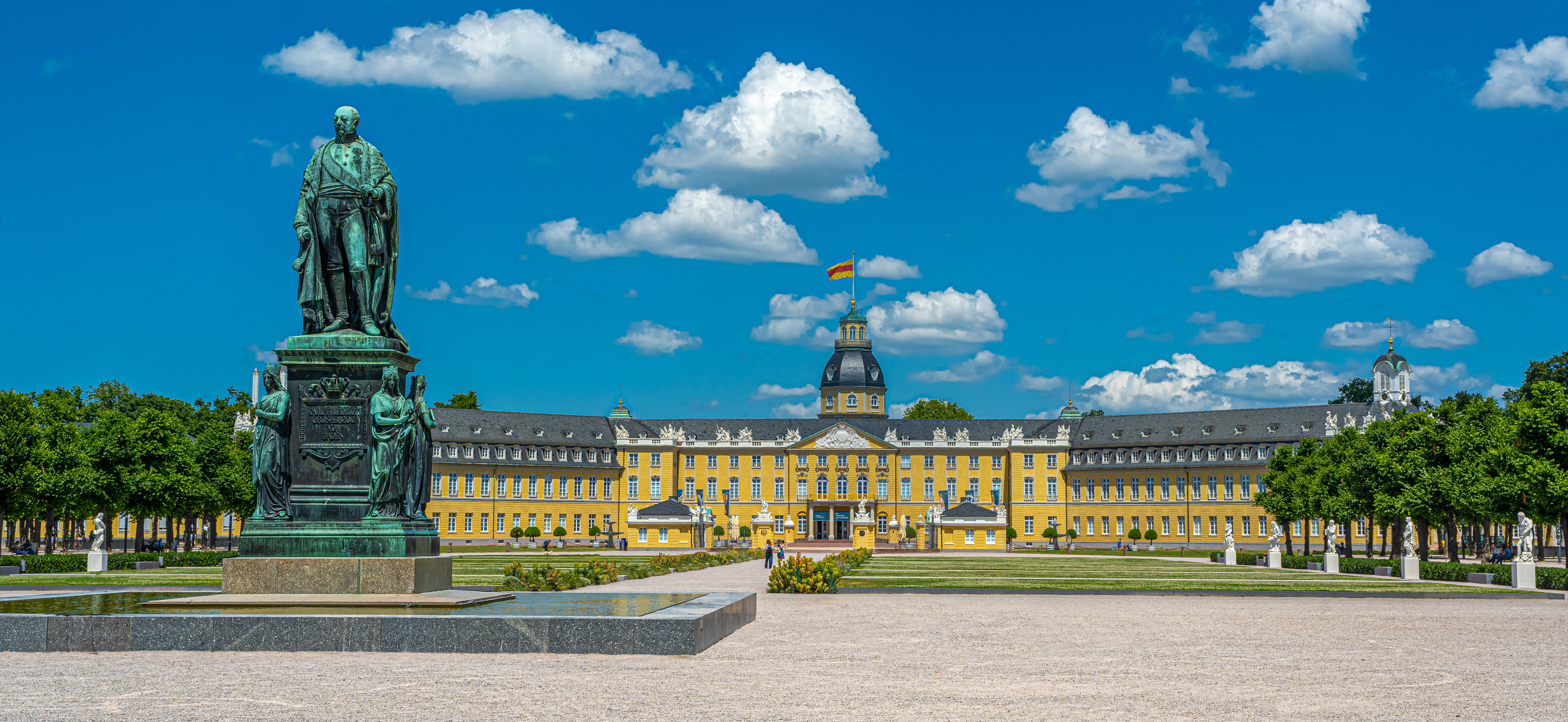 Großes gelbes Schloss mit Turm und Statue im Vordergrund, umgeben von Gartenanlagen.