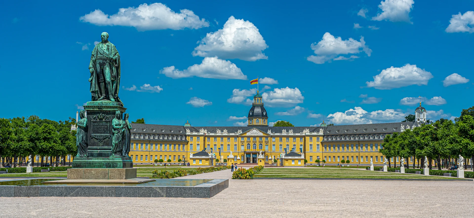 Großes gelbes Schloss mit Turm und Statue im Vordergrund, umgeben von Gartenanlagen.
