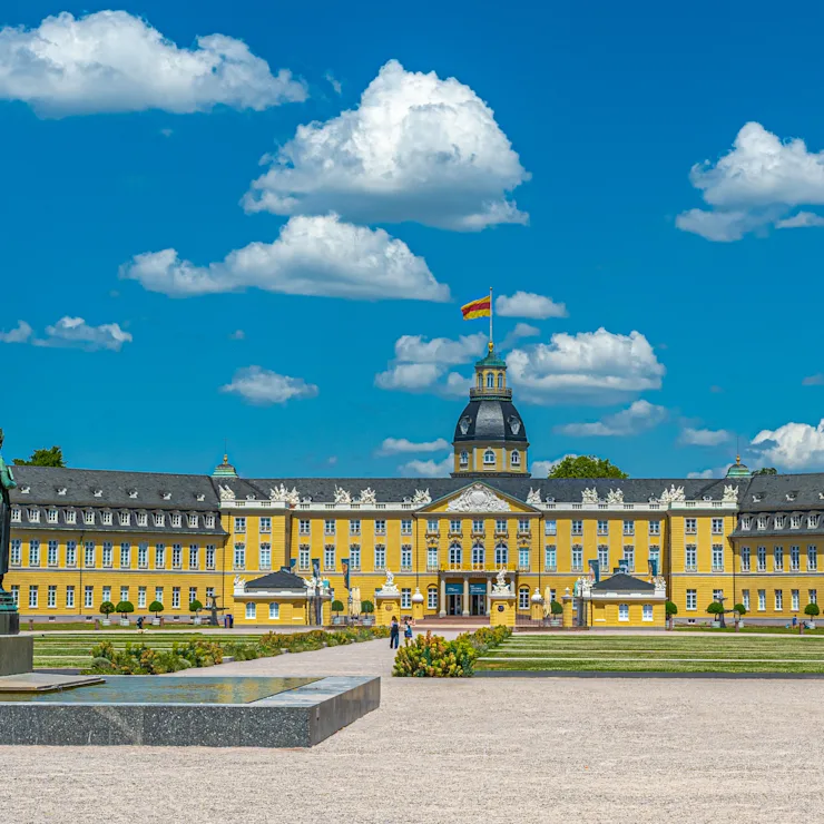 Großes gelbes Schloss mit Turm und Statue im Vordergrund, umgeben von Gartenanlagen.