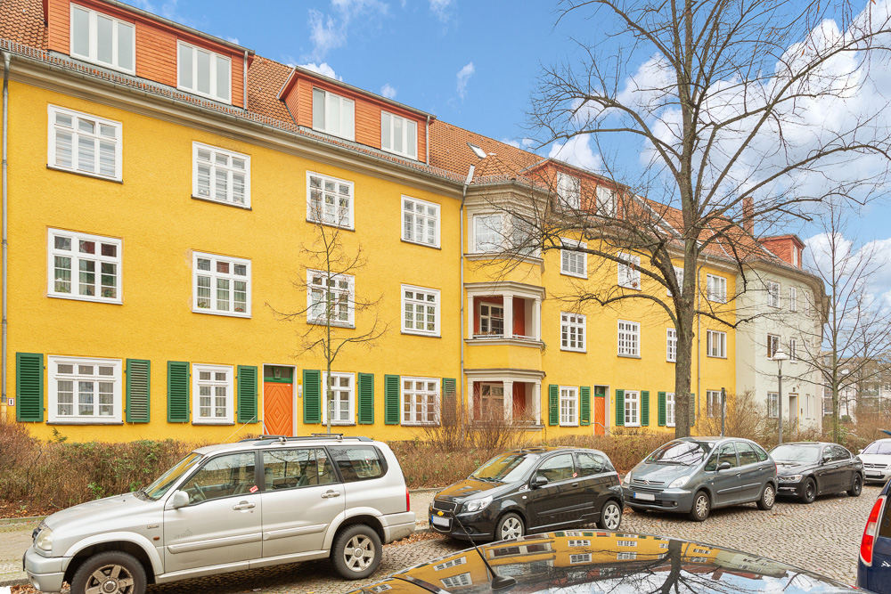 Gelbes Wohngebäude mit grünen Fensterläden, geparkte Autos und Bäume vor dem Haus.