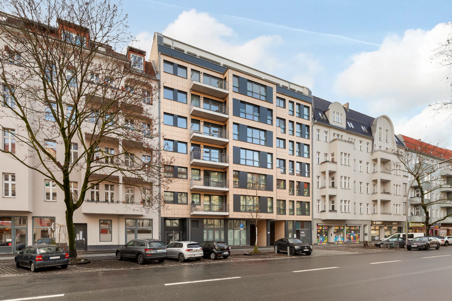 Mehrstöckiges Wohngebäude mit Balkonen, davor parkende Autos an einer Straße.
