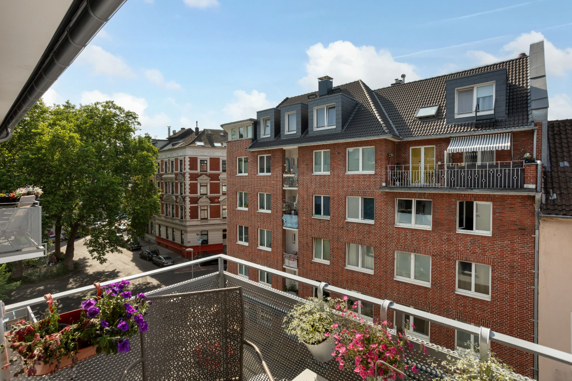 Blick von einem Balkon mit Blumen auf ein rotes Wohnhaus und eine Baumallee.