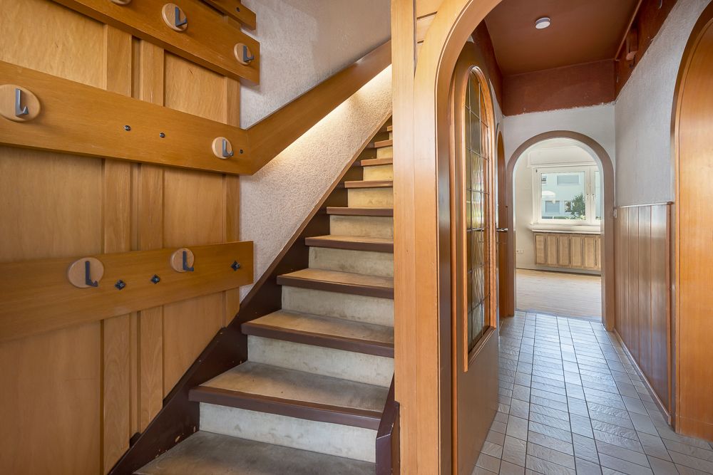 Flur mit Treppe aus Holz und zwei Rundbögen, Fliesenboden und Blick in ein weiteres Zimmer.