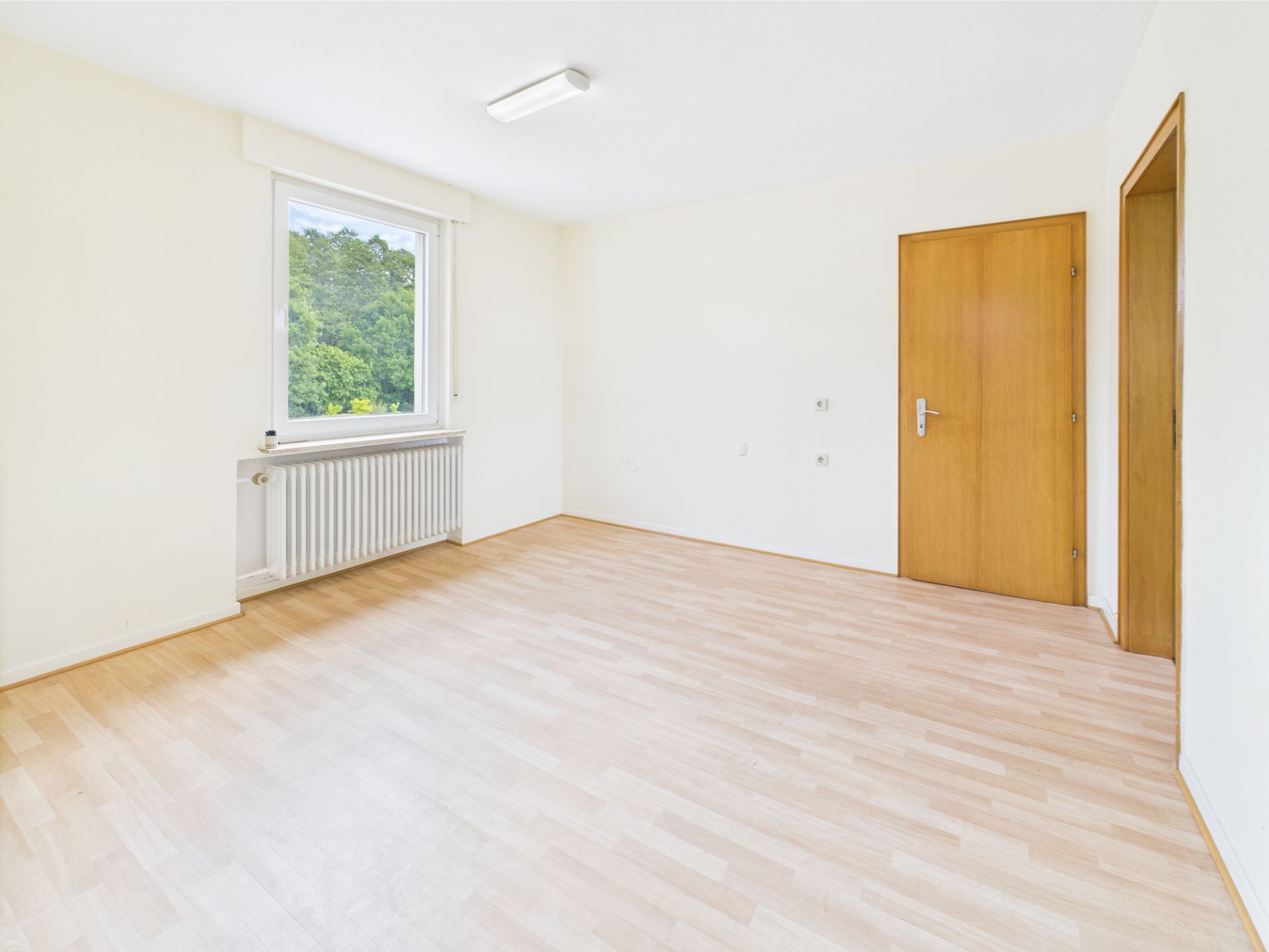 Helles, leeres Zimmer mit Holzfußboden, Fenster, Heizung und zwei Holztüren.