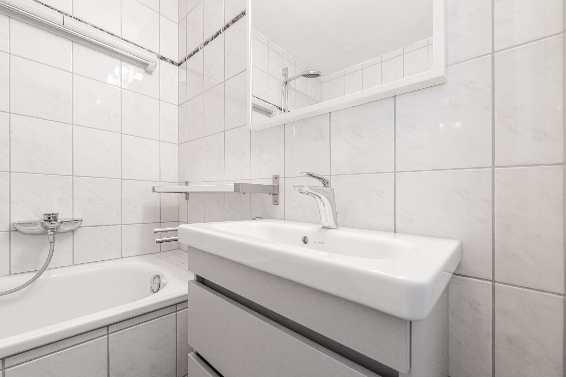 Helles Badezimmer mit Waschbecken, Badewanne und Spiegel, weiße Fliesen an Wand und Boden.