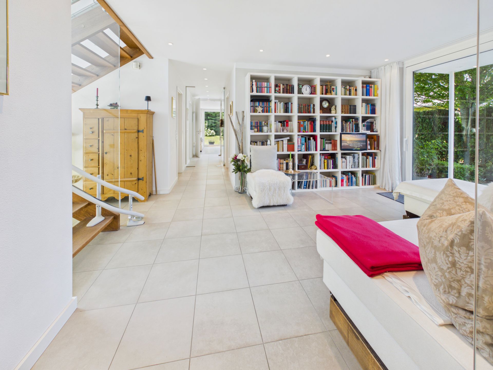 Helles Wohnzimmer mit Bücherregal, Couch, Treppe und großem Fenster zum Garten.