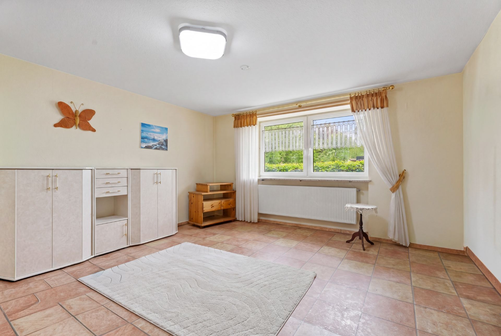 Heller Raum mit Fliesenboden, Teppich, Sideboard, Fenster mit Vorhängen und Wanddeko.