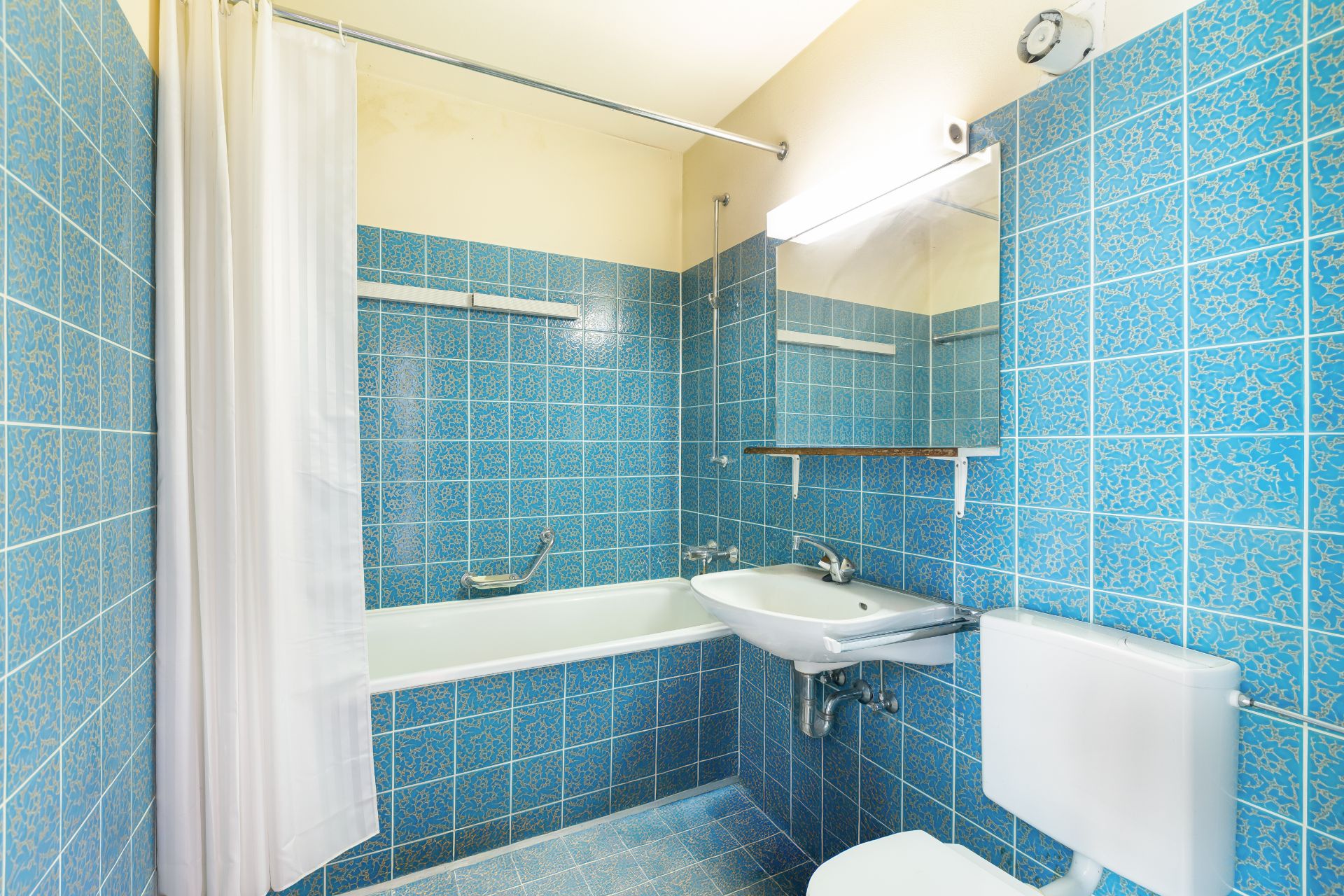 Kleines Badezimmer mit blauen Fliesen, Badewanne, Waschbecken, Spiegel und Toilette.