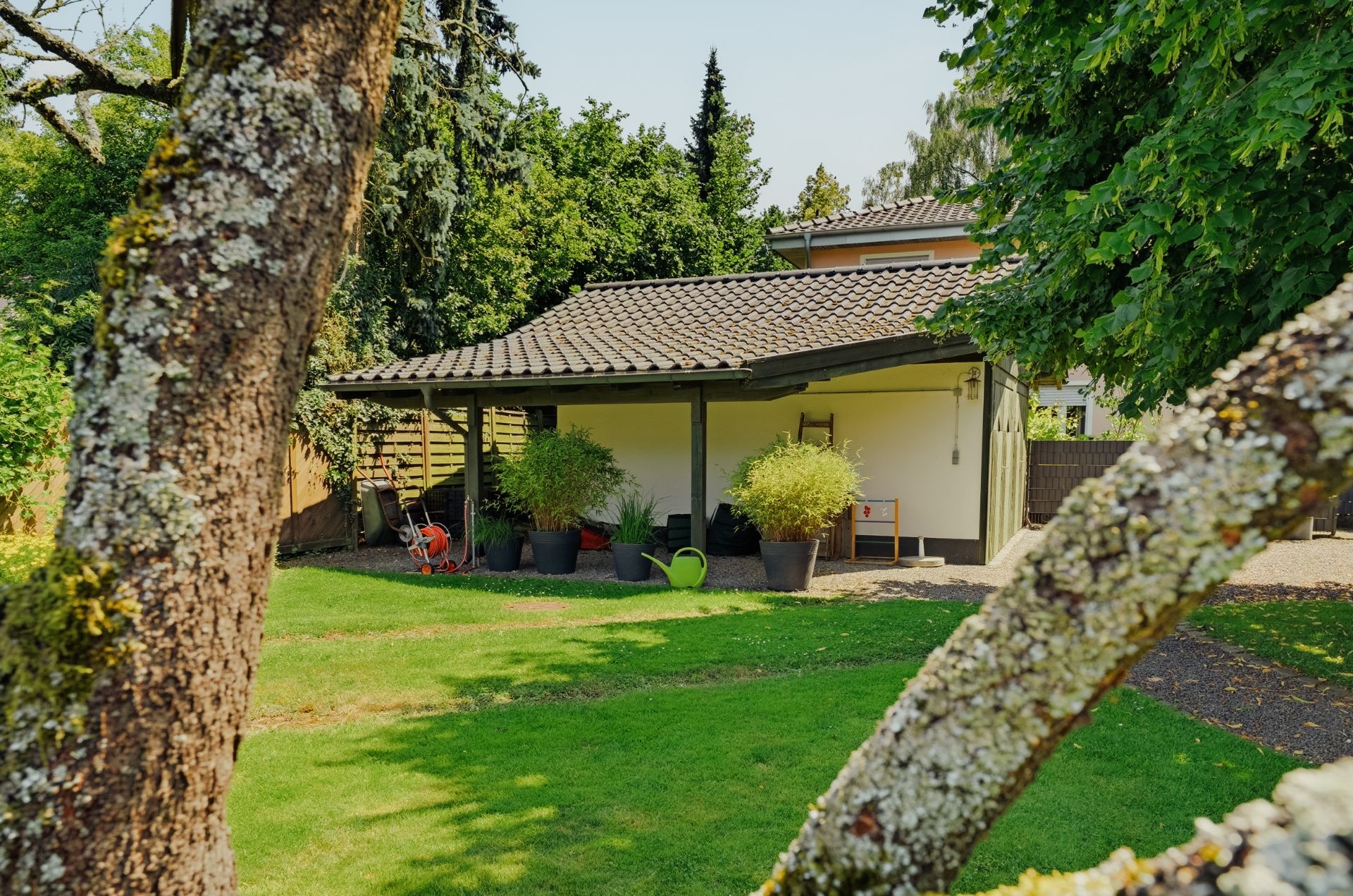 Garten mit Rasen, Bäumen und kleinem überdachten Gebäude mit Blumenkübeln.