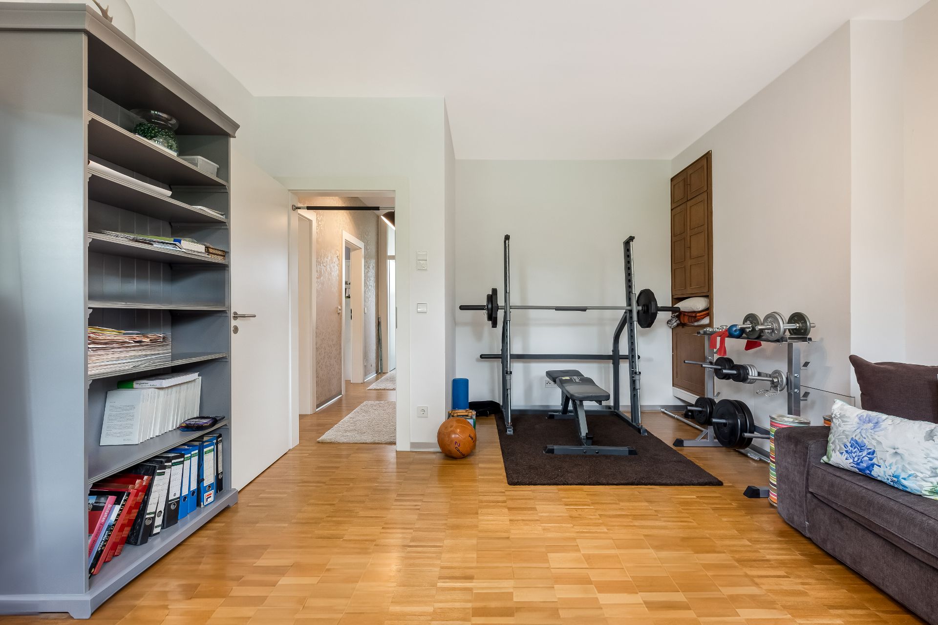 Helles Zimmer mit Hantelbank, Regalen und Couch auf Holzfußboden.