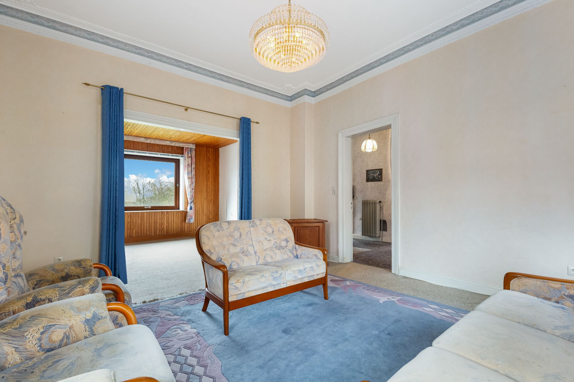 Wohnzimmer mit Sofa, Sesseln, Teppich und Kronleuchter, Fenster mit blauem Vorhang.