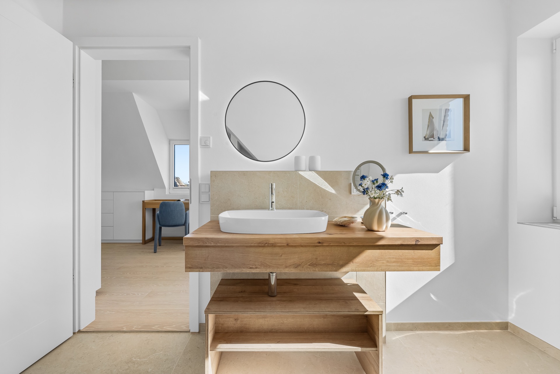 Helles Badezimmer mit Holzw Waschtisch, rundem Spiegel und Vase mit Blumen.
