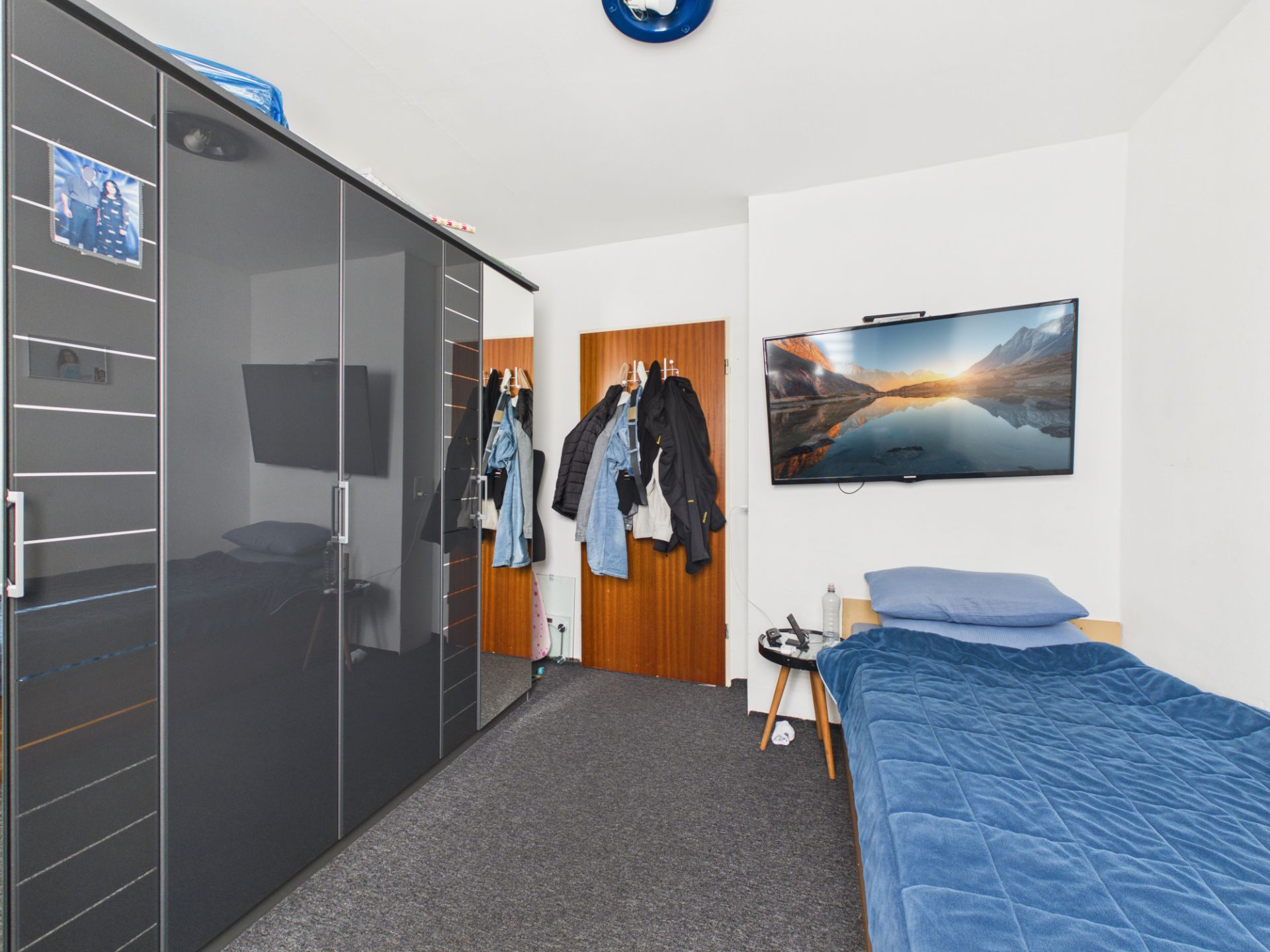 Schlafzimmer mit Einzelbett, schwarzem Kleiderschrank, Fernseher an der Wand und Jacken an der Tür.