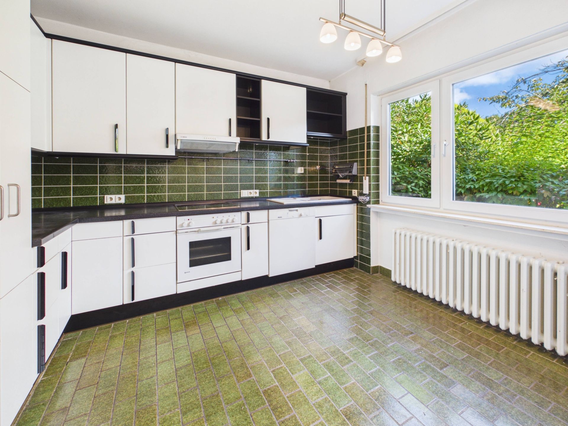 Helle Küche mit weißen Schränken, grünem Fliesenspiegel und großem Fenster mit Blick ins Grüne.