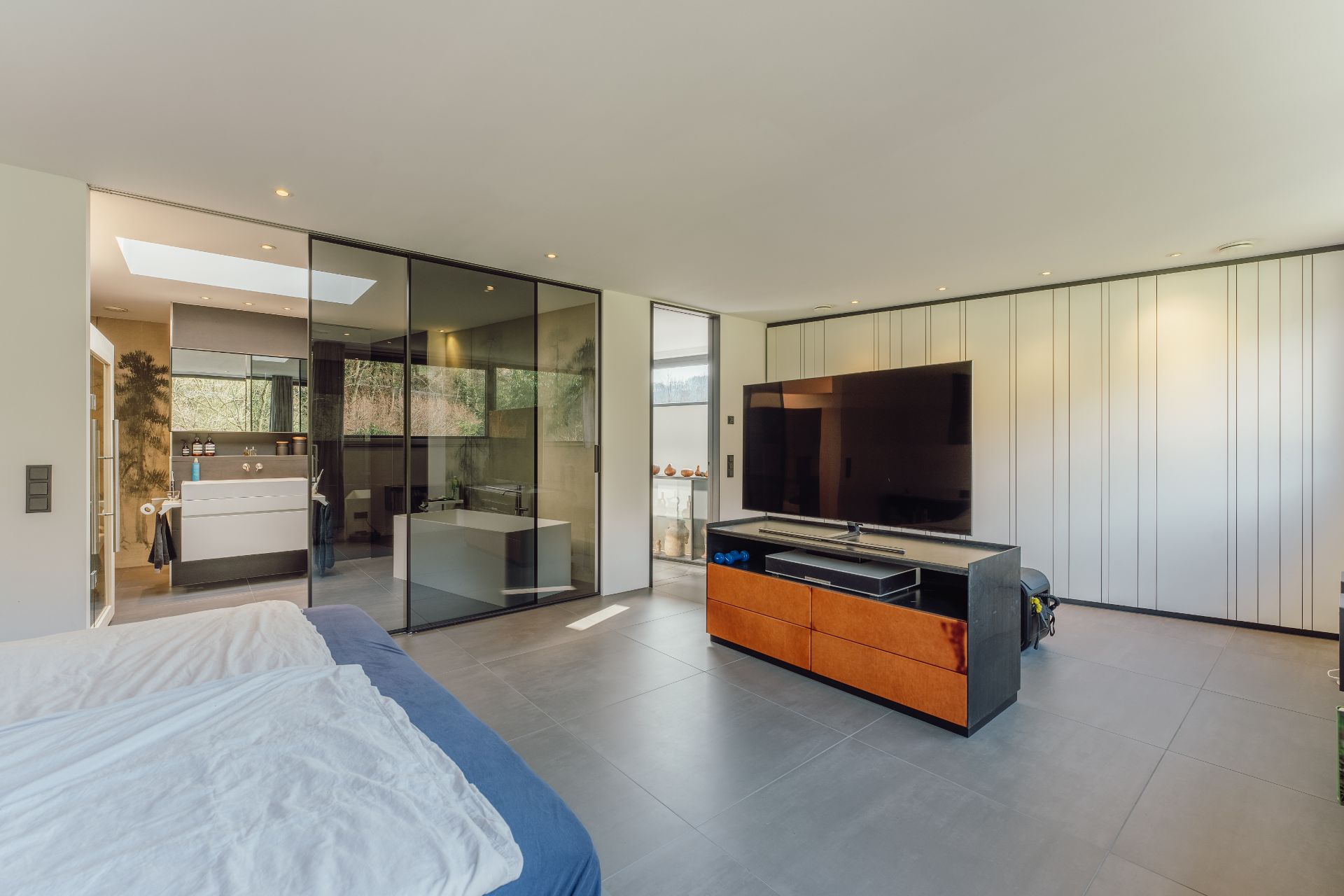 Modernes Schlafzimmer mit großem Fernseher, Doppelbett und Glaswand zum Badezimmer.