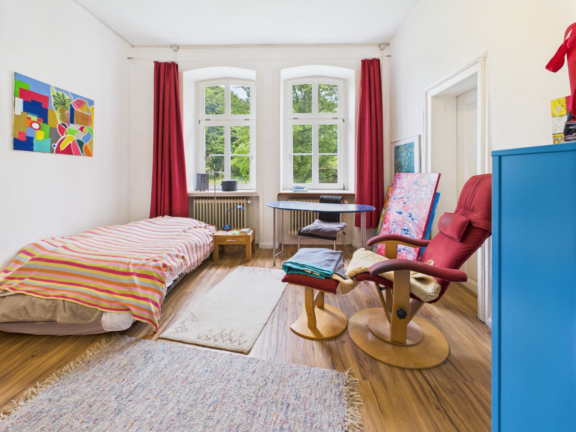 Helles Zimmer mit Bett, Sessel, Schreibtisch, bunten Bildern und roten Vorhängen vor zwei Fenstern.