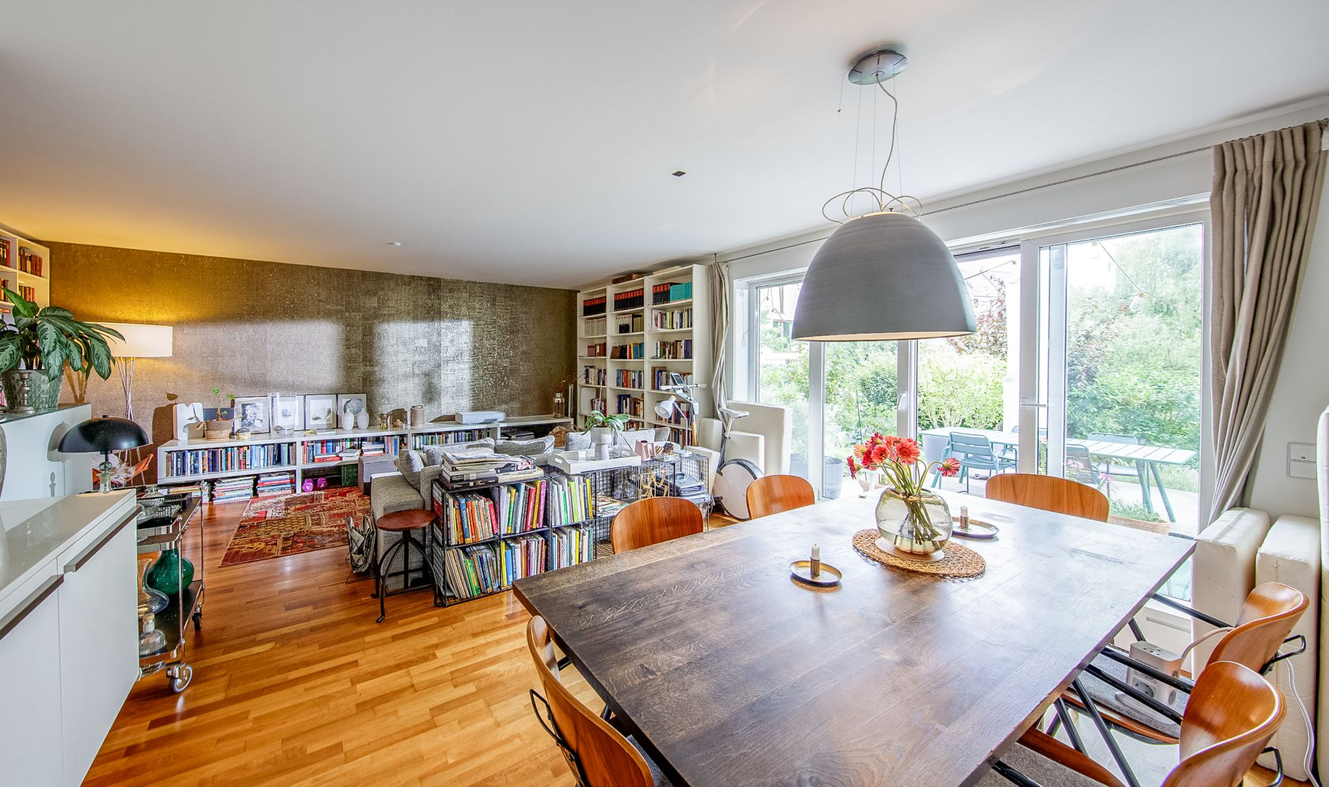 Helles Wohnzimmer mit Holzboden, Esstisch, Bücherregalen und großen Fenstern zum Garten.