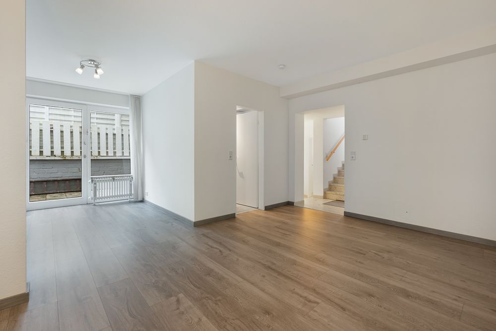 Leeres Wohnzimmer mit Holzfußboden, weißen Wänden und Zugang zu einer Terrasse; Treppe im Hintergrund.