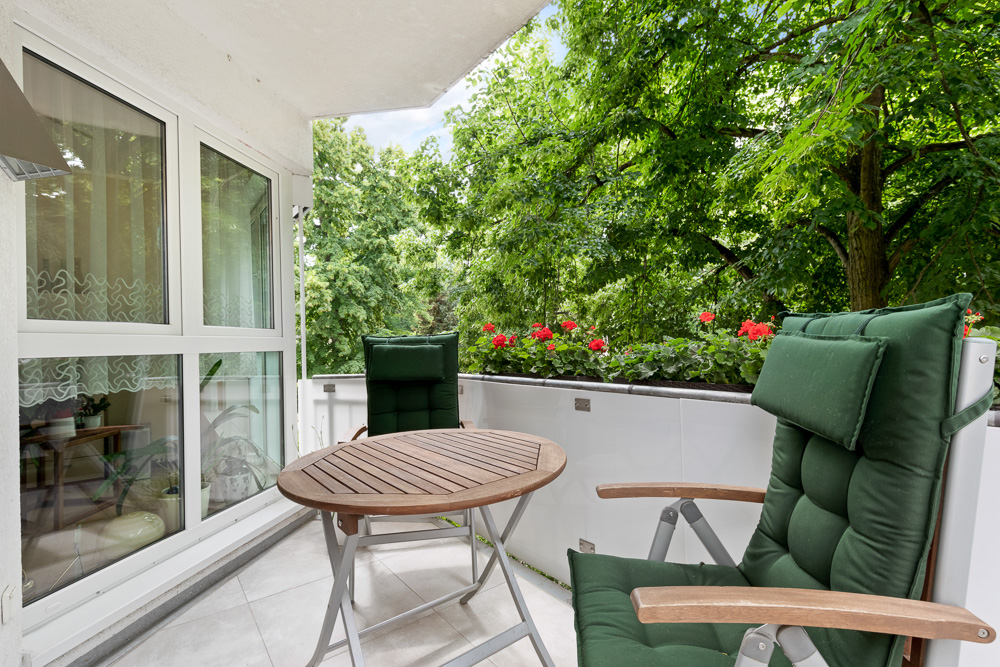 Balkon mit zwei grünen Stühlen, Holztisch und Blumenkasten mit roten Blumen, umgeben von Bäumen.