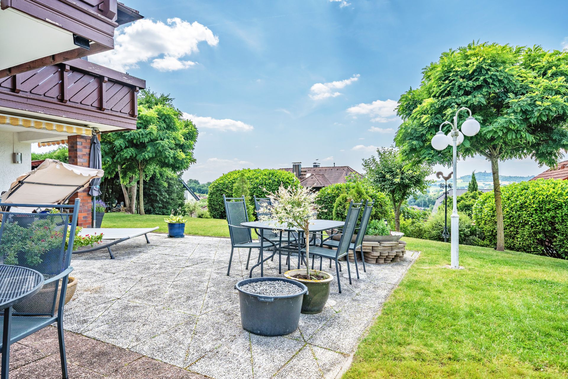 Garten mit Terrasse, Gartentisch, Stühlen, Pflanzen und Bäumen bei sonnigem Wetter.