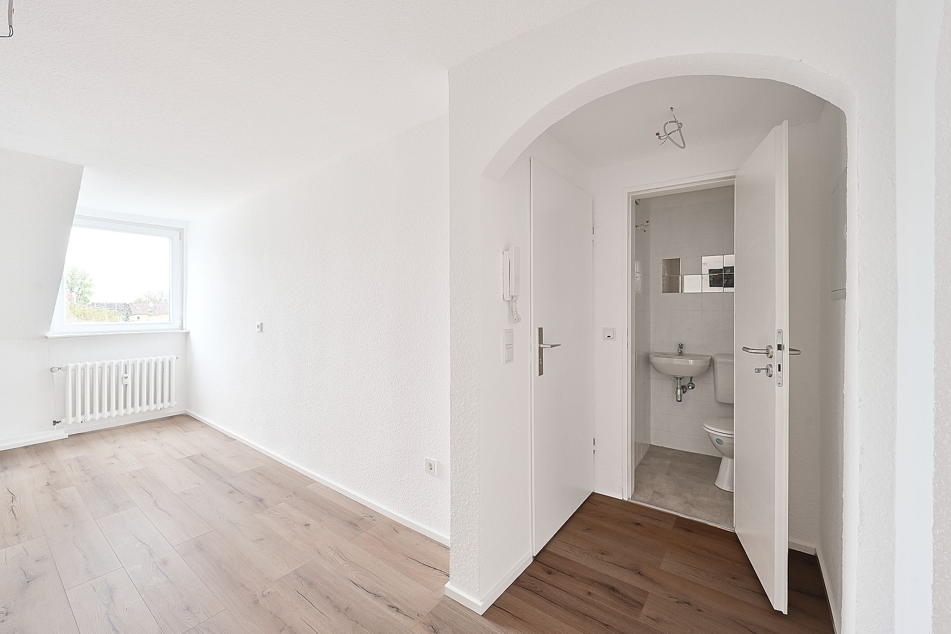 Heller Raum mit Fenster, Holzfußboden und offenem Zugang zu einem kleinen Bad mit Waschbecken und Toilette.