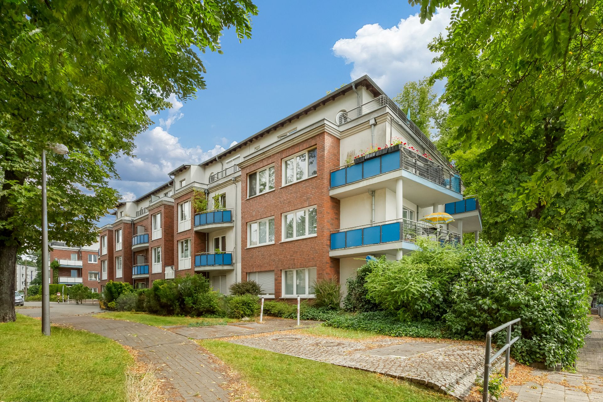 Mehrfamilienhaus mit roten Ziegelwänden, blauen Balkonen und umgeben von Bäumen.
