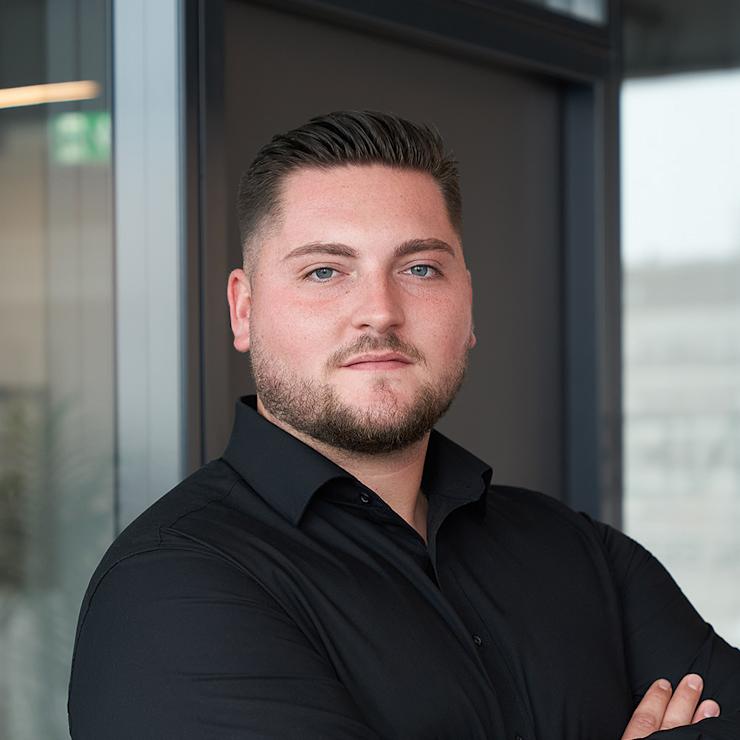 Mann mit Bart und schwarzem Hemd steht mit verschränkten Armen vor Glaswand.