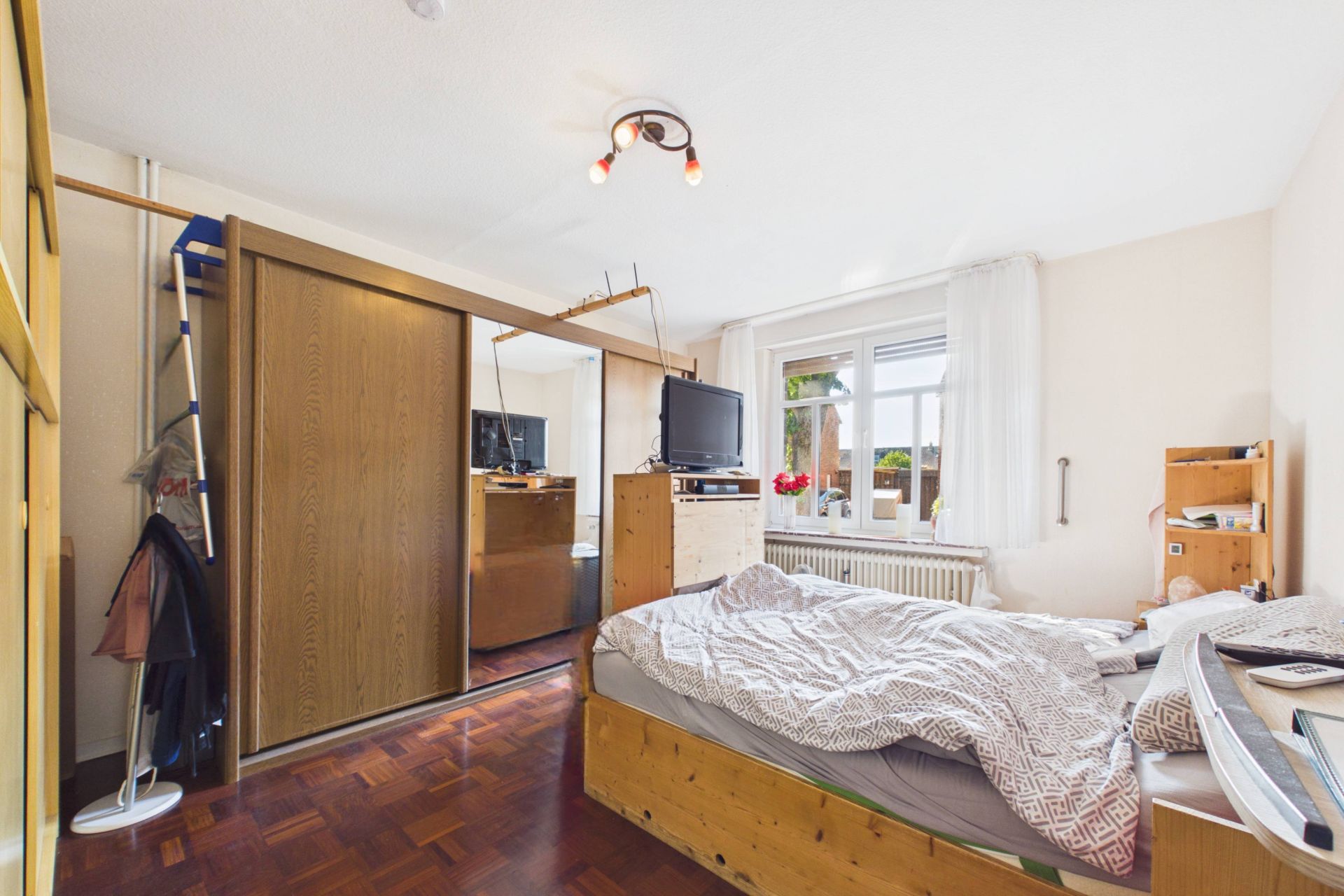 Helles Schlafzimmer mit Holzbett, Kleiderschrank mit Spiegel und Fenster mit weißen Vorhängen.