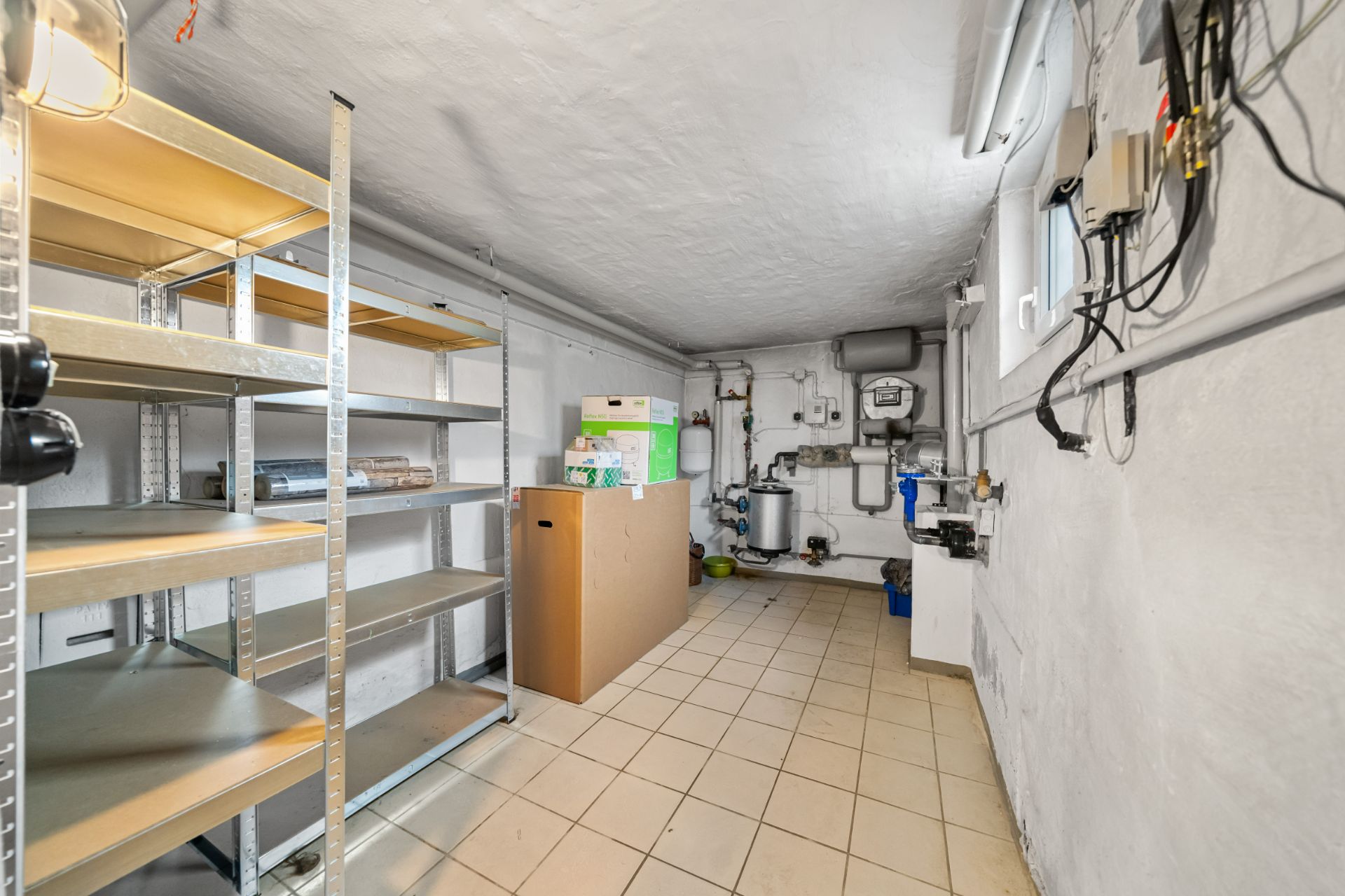 Kellerraum mit leeren Metallregalen, Kisten und Heizanlage an der Wand.
