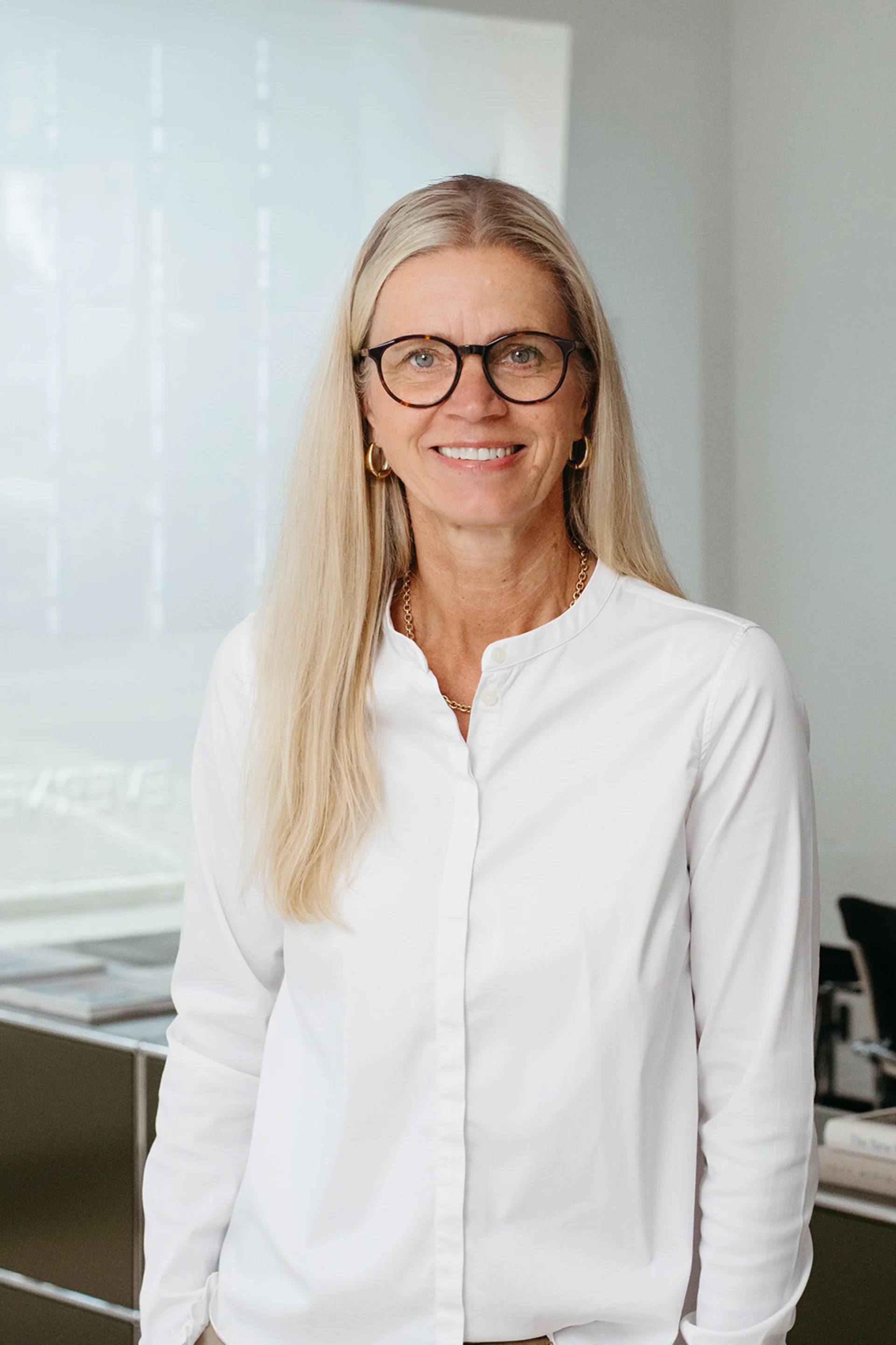 Frau mit langen blonden Haaren und Brille steht in einem modernen Raum.