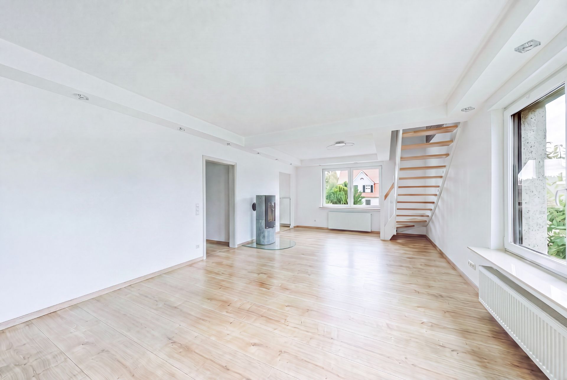 Helles Wohnzimmer mit Holzfußboden, großem Fenster, Kaminofen und Treppe nach oben.