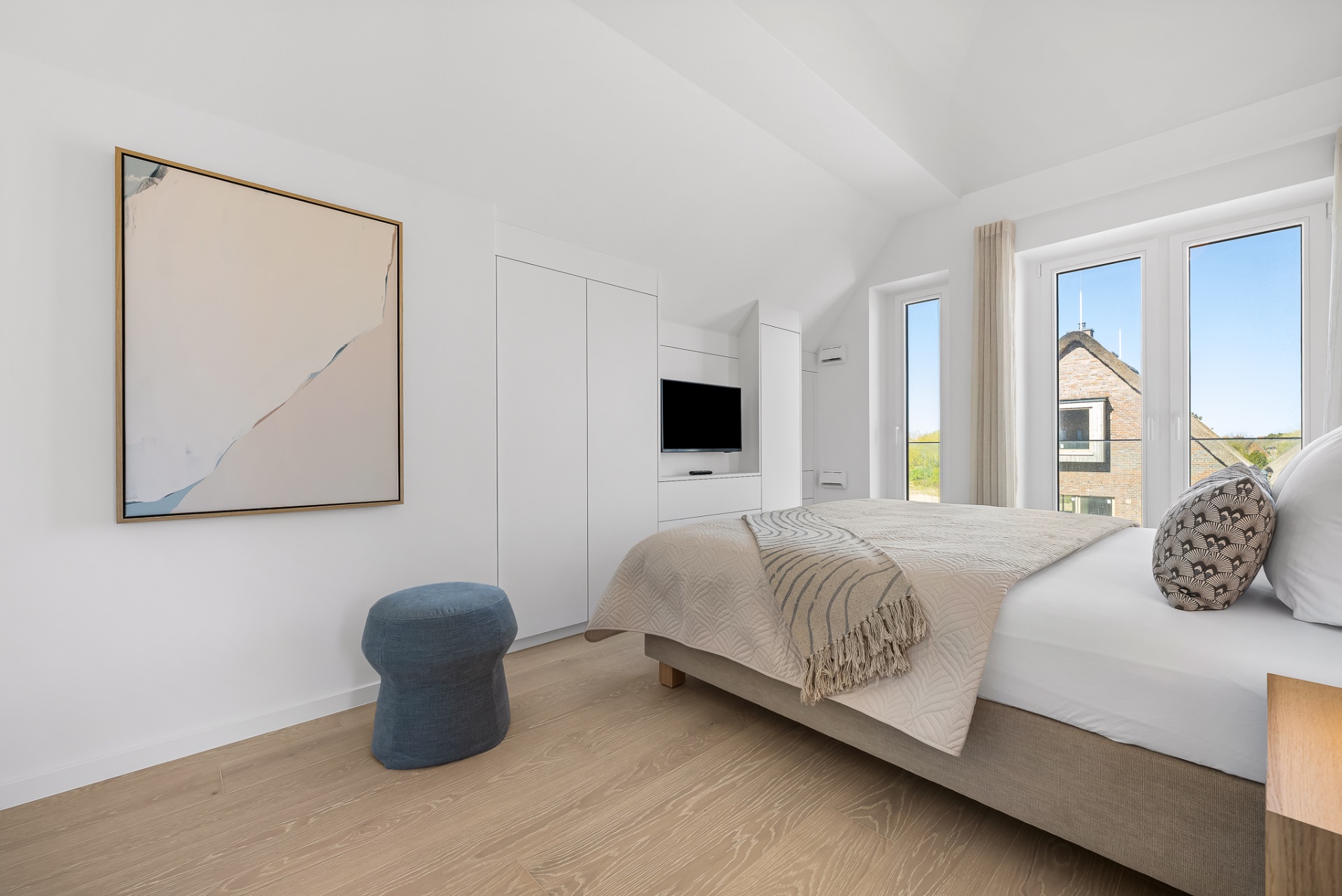 Helles Schlafzimmer mit Doppelbett, großem Fenster, Wandbild und kleinem blauen Hocker.