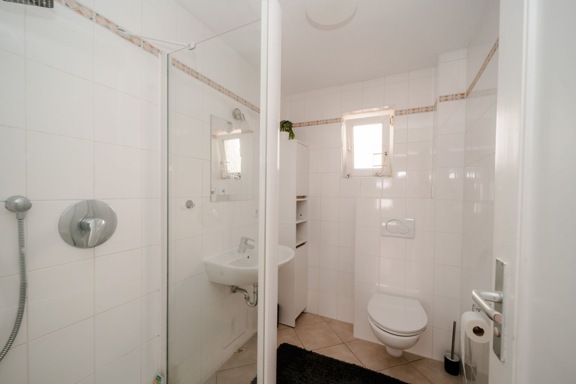 Helles Bad mit Dusche, Waschbecken, WC, kleinem Fenster und weißen Fliesen.