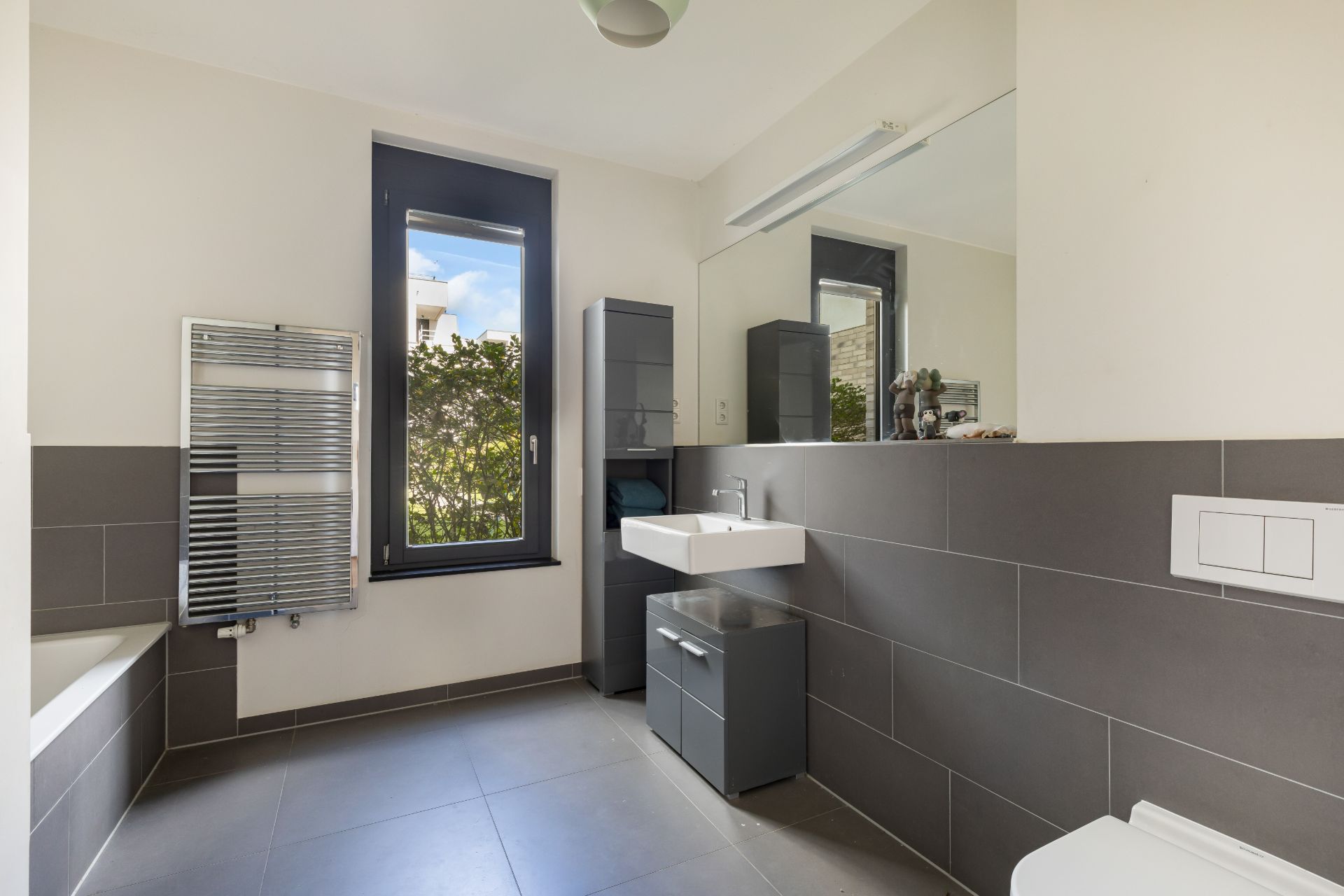 Modernes Badezimmer mit grauen Fliesen, Fenster, Waschbecken, Spiegelschrank und Badewanne.