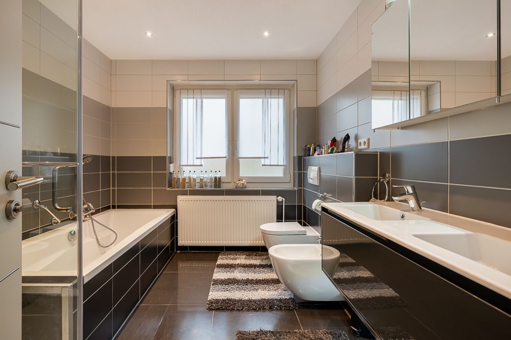 Modernes Badezimmer mit Badewanne, Doppelwaschbecken, WC und Bidet, Fenster und dunklen Fliesen.