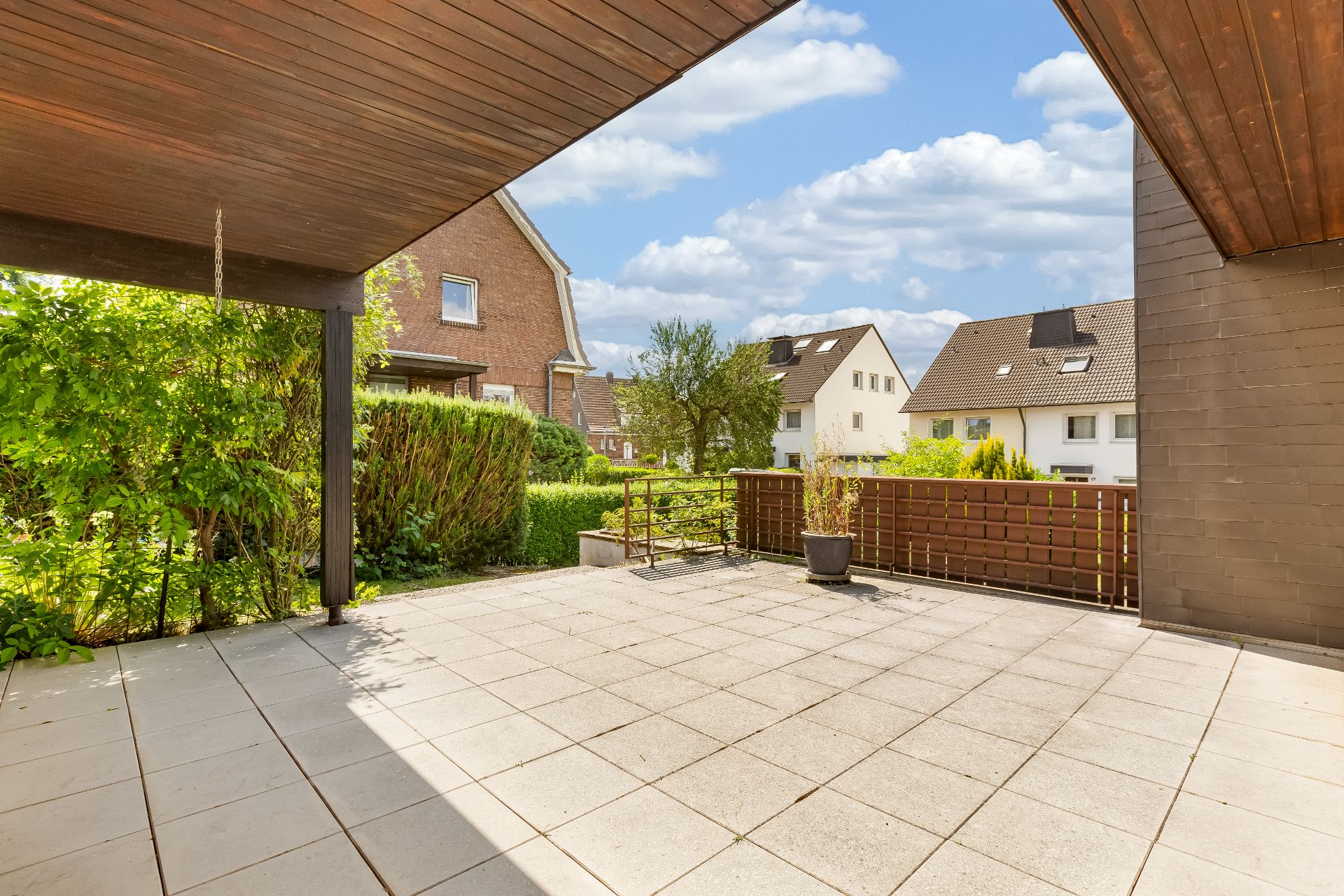 Überdachte Terrasse mit Fliesenboden, Pflanze im Topf und Blick auf Nachbarhäuser und Garten.