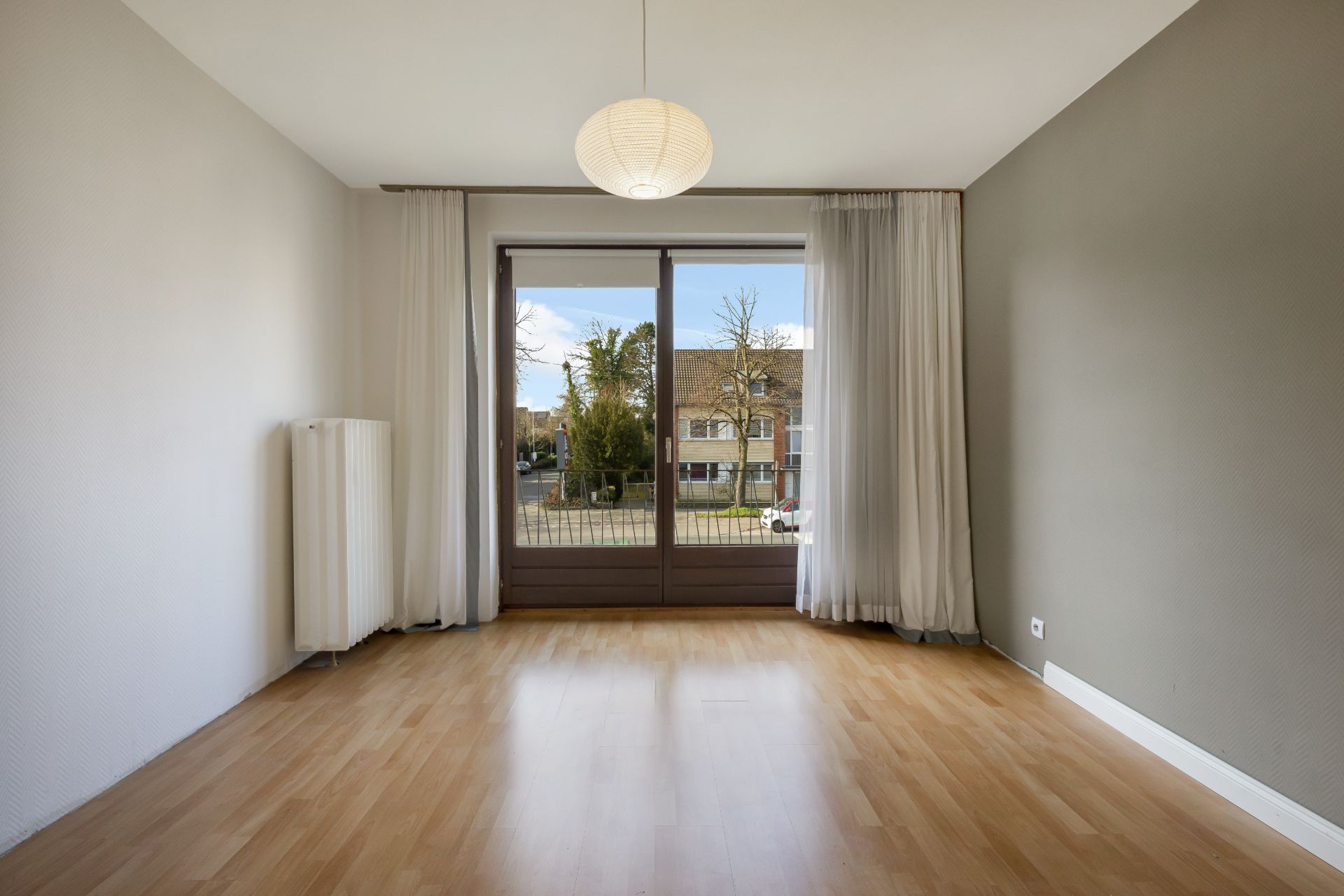 Leeres Zimmer mit Holzfußboden, großen Fenstern und weißen Vorhängen, Blick nach draußen.