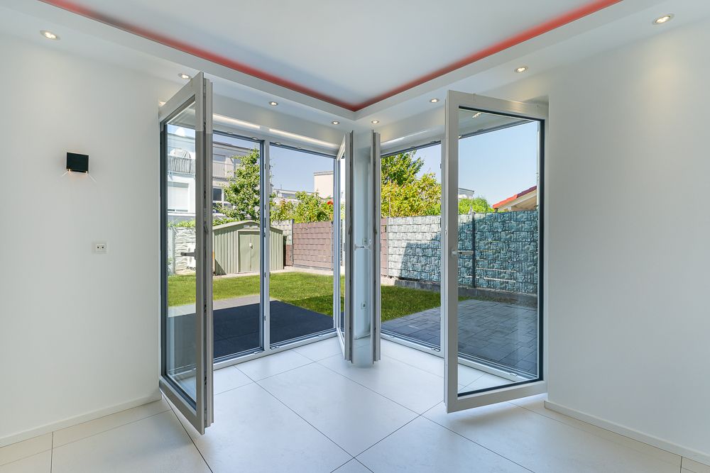 Große, offene Glastüren führen von einem hellen Raum auf eine Terrasse und in einen Garten.