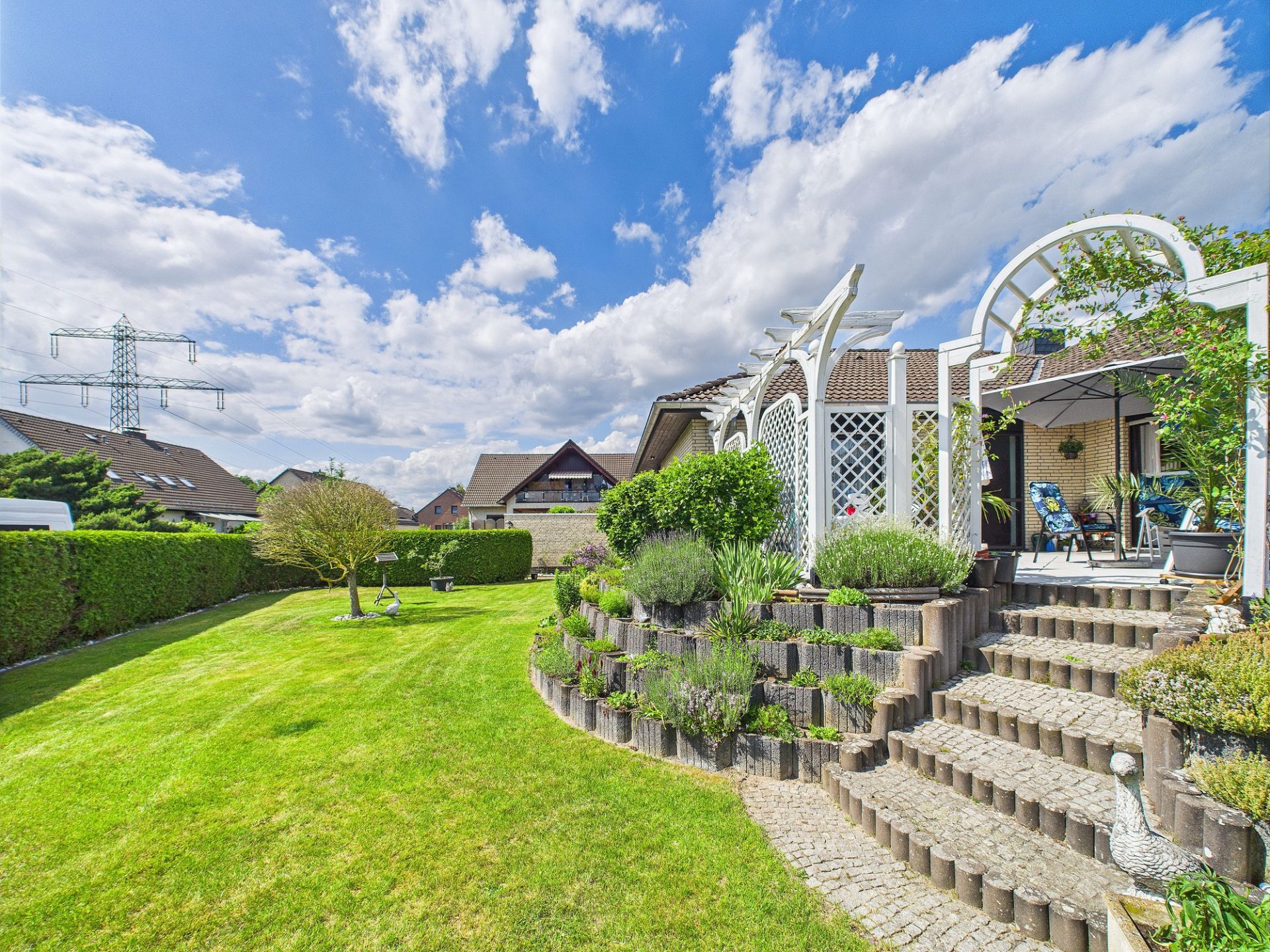 Gepflegter Garten mit Rasen, Baum, Blumenbeeten und einer Terrasse mit Sitzbereich und Pergola.