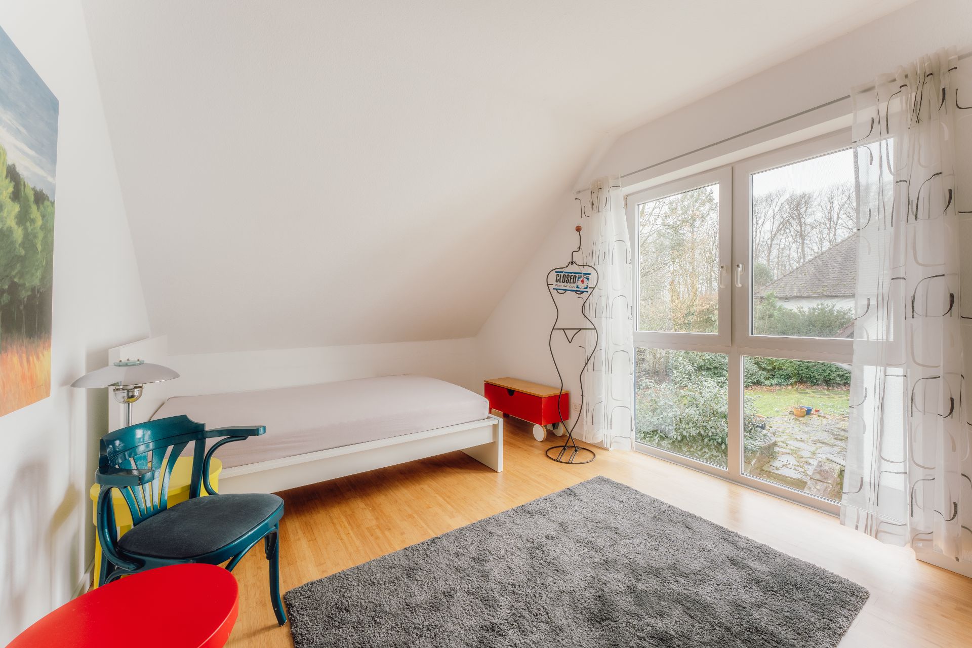 Helles Zimmer mit Einzelbett, Stuhl, Teppich und großem Fenster zum Garten.