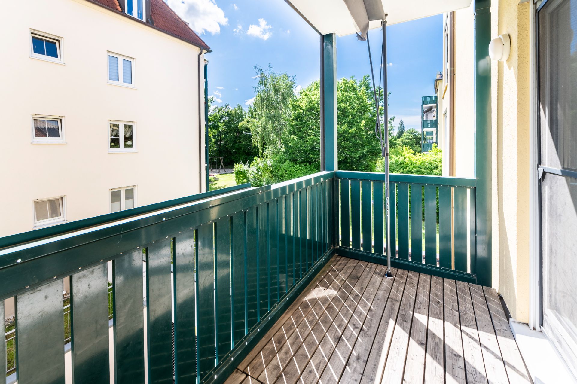 Balkon mit grünem Geländer und Holzboden, Blick auf Bäume und benachbarte Wohngebäude.