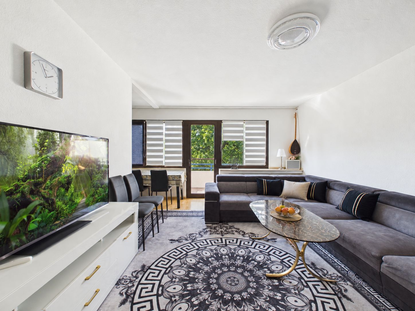 Helles Wohnzimmer mit Ecksofa, Teppich, Couchtisch, Fernseher und großem Fenster.