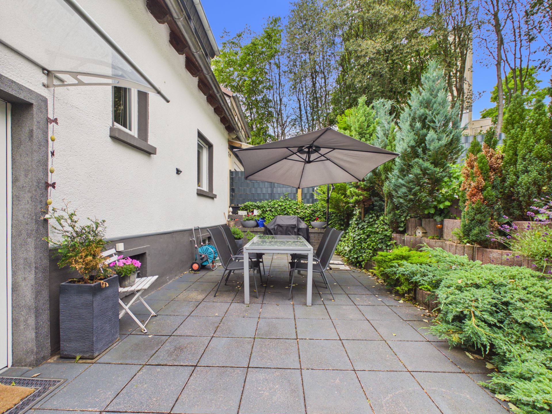 Gepflasterte Terrasse mit Tisch, Stühlen, Sonnenschirm und vielen Pflanzen neben einem Haus.